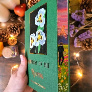 Op de afbeelding: Een groen gebonden boek getiteld Sunrise on the Reaping door Suzanne Collins, met een goudkleurige bloemenillustratie op de voorkant en een beschilderde snede met een persoon die door een veld naar een vulkaan loopt.