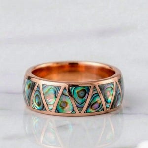 May include: A rose gold band ring featuring a series of triangular inlays made of iridescent abalone shell. The metal frame creates a geometric pattern around the circumference of the ring, showcasing the natural green, blue, and purple hues.