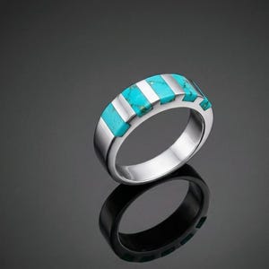 May include: A silver-toned band ring featuring a series of rectangular turquoise stone inlays set vertically along the top half of the outer surface. The metal has a polished, reflective finish, and the turquoise stones display natural blue-green hues.