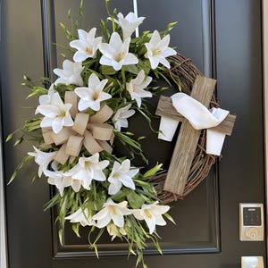 Peut inclure: Une couronne décorative avec des lys blancs, du feuillage vert et un nœud beige. Une croix en bois avec un tissu blanc est attachée à la couronne. La couronne est faite de branches tressées et est affichée sur une porte noire.
