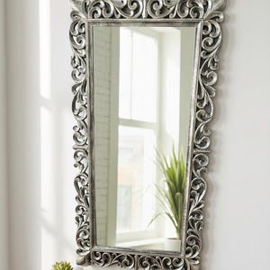 May include: Rectangular silver mirror with ornate, scrolling floral frame. The mirror is mounted on a white wall, reflecting a window and a potted plant. A small succulent in a white pot sits on a wooden shelf below.