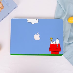 Puede incluir: Funda para portátil azul claro con un dibujo animado de Snoopy descansando en su caseta roja, con Woodstock cerca. La funda tiene una nube blanca y una franja verde en la parte inferior. El logotipo de Apple es visible.