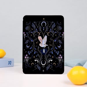 Op de afbeelding: Een rechthoekige e-reader hoes met een zwarte achtergrond en een centrale illustratie van een vliegende kerkuil die een lantaarntje draagt. Het ontwerp bevat symmetrische blauwe en witte botanische ranken, maansikkels, sterren en kleine paddenstoelen.
