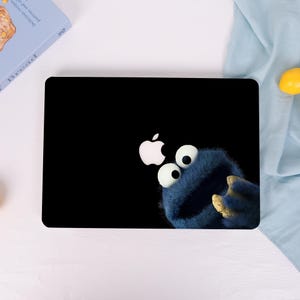 May include: A black laptop skin featuring a graphic of a blue, furry monster with large white eyes and a wide mouth, positioned to appear as if it is reaching toward the white Apple logo centered on the top surface of the device.