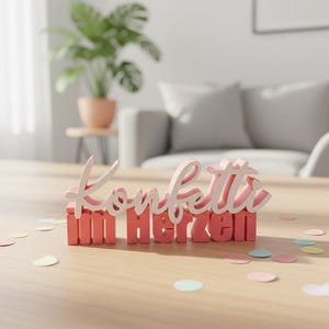 Könnte beinhalten: Ein rosa-weißes Holzschild mit dem deutschen Schriftzug "Konfetti im Herzen" auf einem Holztisch. Der Tisch ist mit Konfetti übersät. Eine Pflanze in einem Terrakotta-Topf und ein graues Sofa befinden sich im Hintergrund.