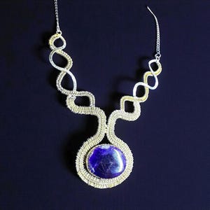 May include: A handcrafted necklace featuring a large, oval, deep blue gemstone pendant. The pendant is encased in a woven silver and gold-toned wire design. The necklace has a silver chain and a unique, openwork design.