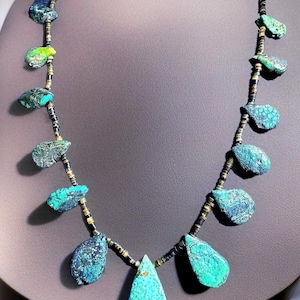 May include: A turquoise and dark blue stone necklace with a teardrop pendant. The necklace features irregularly shaped stones in shades of blue and green, strung on a dark cord with gold-colored accents. The necklace is displayed on a neutral background.