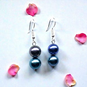 May include: A pair of dangle earrings featuring two iridescent, round beads in shades of blue and gray. The earrings have silver-colored hooks and are displayed with pink rose petals scattered around them.
