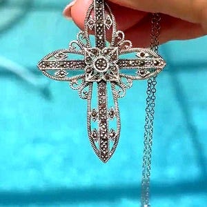 May include: A silver-colored cross pendant with intricate filigree detailing and small, dark gemstones. The cross is suspended from a delicate chain, creating an elegant and ornate necklace.