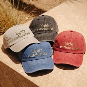 May include: Four baseball caps in different colors: tan, charcoal, blue, and red. Each cap has the phrase "highly caffeinated" embroidered on the front. The caps are arranged on a stone surface.