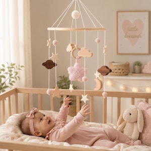 May include: A nursery mobile featuring a circular wooden frame suspended by white strings. It includes hanging elements such as light pink crocheted stars and spheres, smooth wooden stars, clouds, a crescent moon, and a dark brown wooden cloud shape.