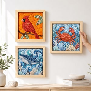 Puede incluir: Tres piezas de arte en tela de forma cuadrada con diseños acolchados. Una representa un cardenal rojo sobre fondo naranja, otra muestra un cangrejo rojo en agua azul y la tercera exhibe un tiburón nadando entre olas azules y conchas marinas.