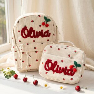 May include: Cream-colored backpack and lunch bag set with a strawberry pattern. The name "Olivia" is embroidered in red script on both items, along with a cherry applique. The backpack has adjustable straps and a front pocket. The lunch bag has a handle.