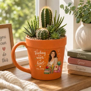 Può includere: Un vaso in ceramica arancione a forma di tazza contenente diverse piante succulente e cactus verdi. La tazza presenta la scritta "Teaching is a work of heart" insieme a un'illustrazione di una persona, libri, un mappamondo e materiale scolastico.