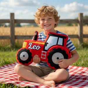 May include: A red tractor-shaped pillow with the name "Leo" in white lettering. The pillow has black wheels and a black smokestack. The tractor is held by a person sitting on a red and white checkered blanket.