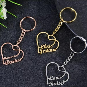 May include: Three heart-shaped keyrings in gold, rose gold, and silver. Each keyring features a double heart design with personalized names or words. The keyrings have a chain and a ring for attaching keys.