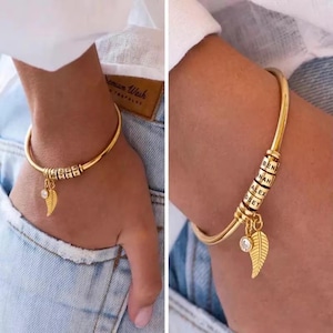 May include: Gold-colored bangle bracelet with personalized name beads and a leaf charm. The bracelet features a delicate leaf pendant and a small, clear gemstone. The names "BEN", "NAH", "ALEX", and "OSEY" are visible on the beads.