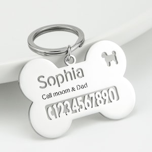 May include: A silver-colored metal pet ID tag shaped like a bone, featuring a circular split ring. The tag is engraved with the name Sophia, the text Call moom & Dad, and the number 1234567890, along with a small cutout silhouette of a dog.