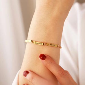 Puede incluir: Una pulsera dorada con placas rectangulares grabadas con los nombres "Eleanor" y "Catelyn". La pulsera se lleva en la muñeca, con una mano y esmalte de uñas rojo visibles. Es una pieza de joyería.
