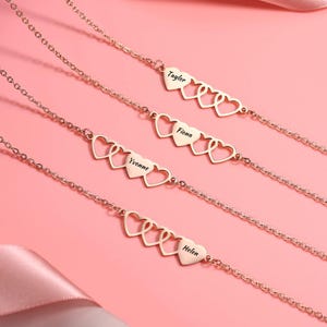 May include: Rose gold heart-shaped bracelets with personalized names. Each bracelet features a chain with three open hearts and a heart-shaped charm with a name engraved. The names include Taylor, Fiona, Yvonne, and Helen.