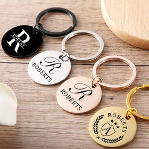 May include: Several personalised keyrings, each engraved with the name "Robert". The keyrings are circular and available in black, silver, and gold. Designs include a monogram and a crest. The keyrings are attached to metal split rings.