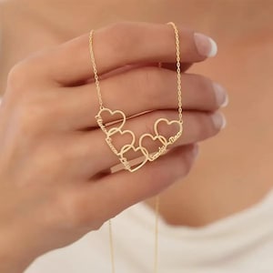 Puede incluir: Un collar dorado con cinco colgantes en forma de corazón, cada uno grabado con un nombre. El collar es sostenido por una mano, resaltando la fina cadena y el diseño personalizado. Un regalo atento.