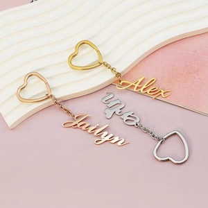 Pode incluir: Três chaveiros personalizados com detalhes em forma de coração. Um é dourado com o nome "Alex", outro é ouro rosa com "Jailyn", e o terceiro é prateado com um nome. Os chaveiros estão em um fundo rosa claro e branco.