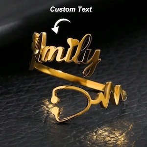 Könnte beinhalten: Ein goldfarbener Ring mit dem Namen "Amily" in Schreibschrift. Der Ring hat ein Stethoskop-Design und den Text "Custom Text" über dem Ring. Der Ring befindet sich auf einer dunklen Oberfläche.
