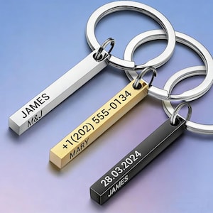 May include: Three personalised keyrings with rectangular bars in silver, gold, and black. The silver keyring has "JAMES" and "M&J" engraved. The gold keyring displays a phone number and "MARY". The black keyring shows a date and "JAMES". All have silver keyrings.