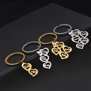 May include: A collection of personalized heart-shaped keychains in gold and silver tones. Each keychain features names like "Lay," "Lily," and "Maly" engraved on the heart pendants. The keyrings are displayed on a dark surface.