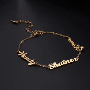 May include: A gold-coloured bracelet featuring the names "Wendy", "Shaitnez", and "Ama" in a cursive font. The bracelet has a fine chain and a clasp. The background is black.