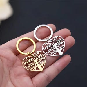 Op de afbeelding: Hartvormige sleutelhangers met een boomontwerp, elk gegraveerd met namen. Eén sleutelhanger is goudkleurig, de andere is zilverkleurig. De namen "Tony", "Jessica", "Daniel", "Sofia", "Andy" en "Cherry" zijn zichtbaar.