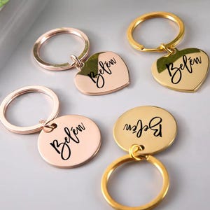 May include: Gold and rose gold keyrings with pendants engraved with the name "Belén" in cursive script. The pendants are round and heart-shaped. The keyrings are gold and rose gold.