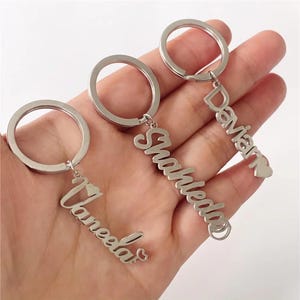 Puede incluir: Tres llaveros plateados con nombres personalizados: "Vaneela", "Shahleena" y "Damian". Cada llavero presenta un colgante con el nombre en cursiva y un corazón. Los llaveros son circulares y están unidos a los colgantes.