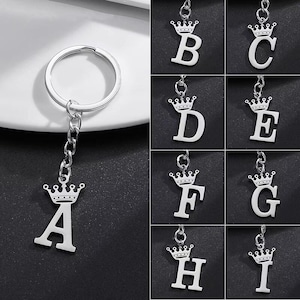 Könnte beinhalten: Silberfarbene Initialen-Schlüsselanhänger mit Kronen-Design. Der Buchstabe "A" ist an einem Schlüsselring befestigt, während die Buchstaben "B" bis "I" separat angezeigt werden. Jeder Buchstabe ist mit einer Krone versehen und mit einer Kette verbunden.