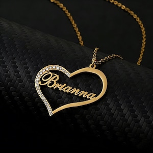 May include: A gold-colored heart-shaped pendant necklace with the name "Brianna" in cursive script. The necklace features a chain and a border of small, clear crystals along the left side of the heart.