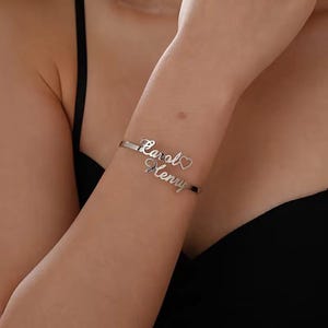 Puede incluir: Una pulsera plateada con los nombres "Carol" y "Henry" en escritura cursiva. Un pequeño corazón está entre los nombres. La pulsera se lleva en una muñeca.