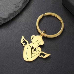 Puede incluir: Llavero dorado que representa a una madre ángel sosteniendo a un bebé ángel, ambos con alas. El llavero tiene un anillo redondo para llaves. El diseño es una silueta, con la madre y el bebé en un abrazo amoroso.