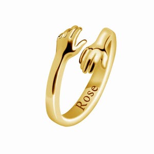 Puede incluir: Un anillo dorado con dos manos que se abrazan. La palabra "Rose" está grabada en la banda. Las manos son detalladas, con dedos visibles y un acabado liso y pulido. El anillo está sobre un fondo blanco.