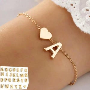 Puede incluir: Pulsera dorada con una delicada cadena, con un dije de corazón y la letra "A". La pulsera se muestra en una muñeca, con una pequeña exhibición de otros dijes de letras en la esquina inferior izquierda. Un accesorio elegante y personalizado.