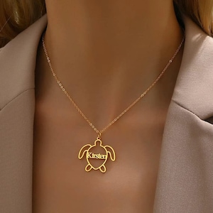Könnte beinhalten: Eine goldfarbene Halskette mit einem Schildkrötenanhänger. Der Anhänger hat den Namen "Kirsten" in der Mitte ausgeschnitten. Die Halskette wird um den Hals getragen.