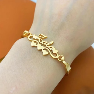 May include: A gold-colored bracelet with the name "Lala" in a cursive font, accented with heart shapes. The bracelet is a bangle style, with a clasp closure. The jewelry is displayed on a person's wrist.