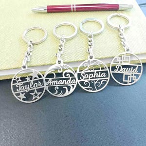 Puede incluir: Cuatro llaveros plateados con nombres personalizados: Taylor, Amanda, Sophia y David. Cada llavero presenta un diseño circular con recortes decorativos y una cadena. Un bolígrafo rojo y plateado descansa en el fondo.