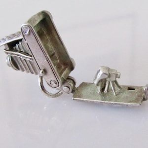 May include: A silver miniature camera charm with an open compartment. The camera features a detailed bellows design and a hinged opening, revealing a small, detailed scene inside. The charm is likely made of metal and has a vintage aesthetic.