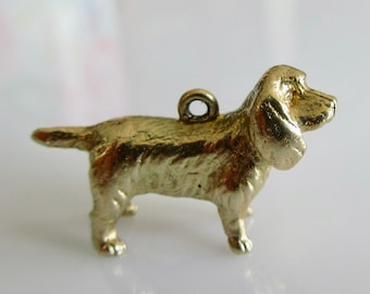 May include: A small, gold-colored dog charm with a loop for hanging. The charm is a detailed miniature sculpture of a dog, with visible features like ears, tail, and legs. The dog is standing, and the overall design is simple and elegant.