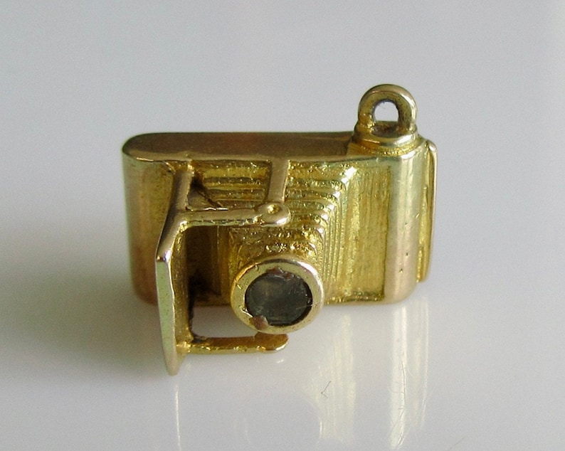 May include: A gold-toned charm in the shape of an old-fashioned camera. The camera has a round lens and a detailed design.