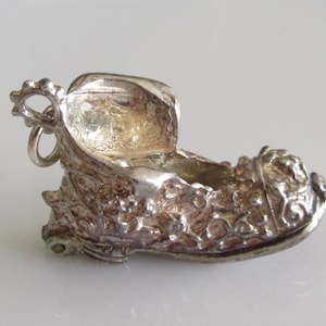 May include: A detailed silver-toned charm in the shape of a shoe. The shoe charm has an open top and intricate floral and scrollwork designs. A small loop is attached for hanging.