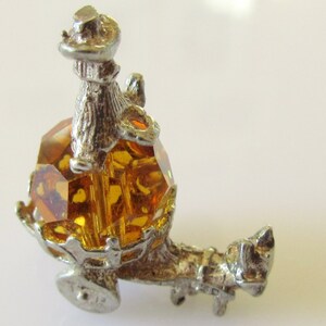 May include: A silver charm featuring a small figure of a person riding in a carriage with a large amber-colored crystal. The carriage has two wheels and a horse pulling it.