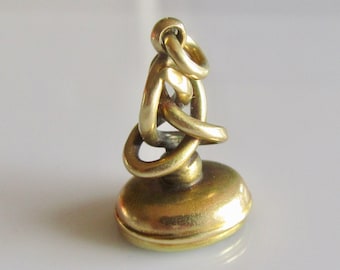 May include: A gold-toned metal seal with an oval base and a decorative, intertwined chain-like top. The seal is designed for attaching to a chain or fob.