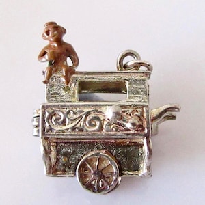 May include: A vintage silver-toned charm depicting a miniature organ grinder's cart. A small brown monkey figure sits on top. The cart has decorative scrollwork and a small wheel.